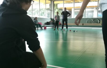 Cerca de cinco pessoas na foto. durante um jogo de boccia. Ao centro da foto vê-se um conjunto de bolas azuis, vermelhas e uma branca.