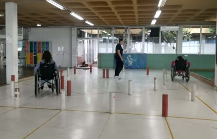 Espaço amplo, com pinos brancos e vermelhos a criarem um percurso. Duas pessoas, em cadeiras de rodas, fazem o percurso e uma terceira pessoa assiste.