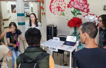 Cerce de cinco pessoas estão à volta de uma mesa onde está um computador e uma série de papéis. A parede, no fundo, tem diferentes cravos colados.