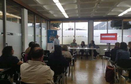 Um homem fala em frente a uma sala de aula onde várias pessoas estão sentadas. Ao lado, há um monitor que exibe o texto 