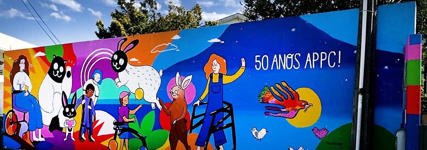Mural colorido com inúmeras imagens pintado em parede exterior. Atrás, ao fundo, algumas árvores.