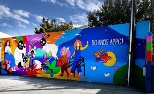 Mural colorido com inúmeras imagens pintado em parede exterior. Atrás, ao fundo, algumas árvores.