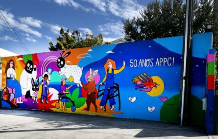 Mural colorido com inúmeras imagens pintado em parede exterior. Atrás, ao fundo, algumas árvores.