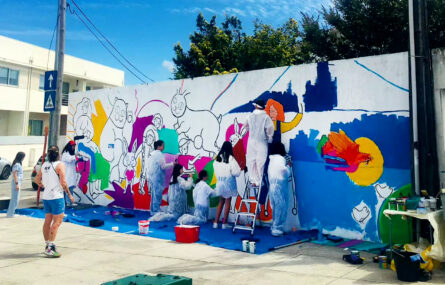 Cerca de uma dezena de pessoas junto de uma parede exterior parcialmente desenhada e a ser pintada. Materiais de pintura estão espalhados por todo o lado. Atrás, ao fundo, algumas árvores e edifícios.