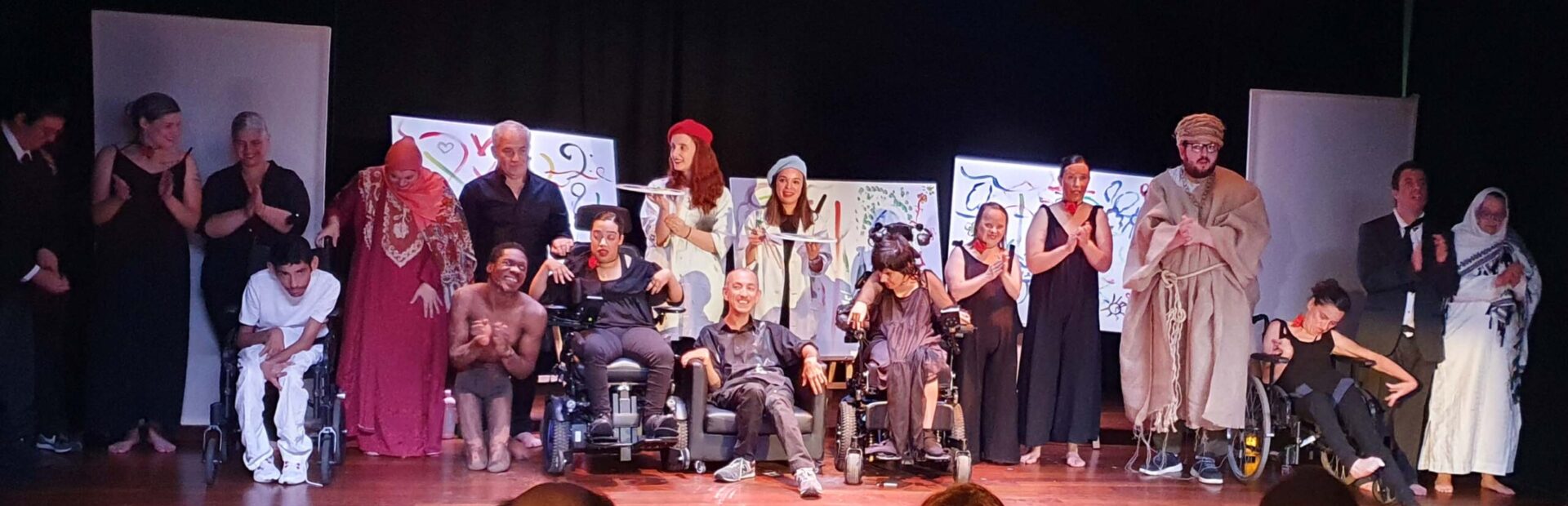 Quase duas dezenas de atores e atrizes alinhados em palco. Em primeiro plano, de costas, algumas pessoas aplaudem.