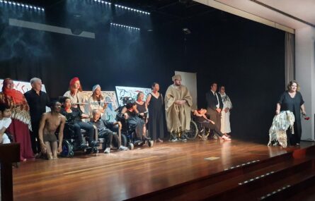 Fotografia de considerável parte de elementos da companhia de teatro em palco. Do lado direito, ligeiramente afastada, aparece uma outra pessoa com um adereço da peça na mão direita e com cravos na mão esquerda.