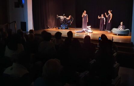 Fotografia de palco e de parte do público a assistir a peça de teatro. Em palco estão seis elementos da companhia de teatro da APPC.