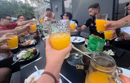 Cerca de uma dezena de pessoas faz um brinde (com copos cheios do que aparenta ser sumo de laranja). Mesa com alguns produtos alimentares.
