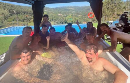 Mais de uma dezena de pessoas naquilo que aparenta ser uma pequena piscina exterior (ou jacuzzi). AO fundo é visível uma piscina exterior, espaços verdes, árvores e montes.