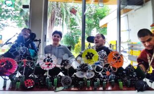Quatro pessoas, sorridentes, junto de um trabalho artesanal elaborado com diversos materiais indistintos. Ao fundo, pelas portadas em vidro, vê-se um espaço ajardinado.