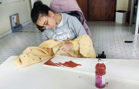 Uma pessoa, junto de uma mesa, utiliza um pincel para pintar em tons de castanho o que aparenta ser um quadro. Na mesa está ainda pousado um recipiente com tinta castanha.