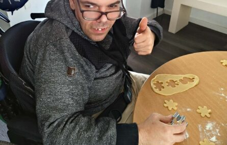 Uma pessoa utiliza peça em metal para cortar pedaços de massa em formato de biscoitos. A pessoa em questão olha para o fotógrafo e faz um sinal de “fixe” com o dedo polegar da mão esquerda.