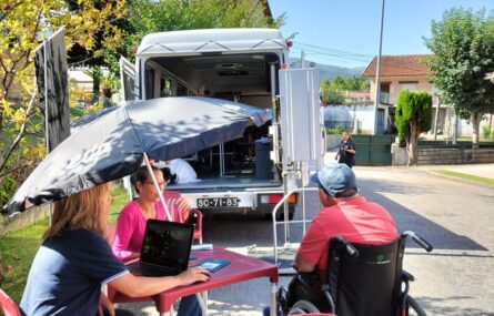Três pessoas sentadas à volta de uma mesa de esplanada. Uma das pessoas está a utilizar um computador. Uma das pessoas é utilizador de cadeira de rodas manual. Ao fundo, vê-se o autocarro adaptado. 