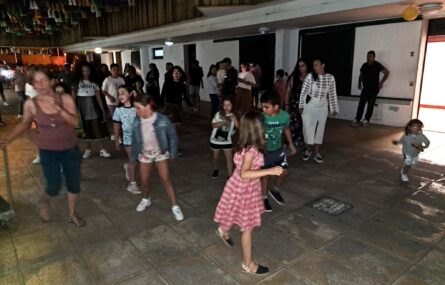 Várias dezenas de pessoas – crianças, jovens e adultos – a dançar em espaço exterior. Fotografia noturna e com pouca visibilidade.