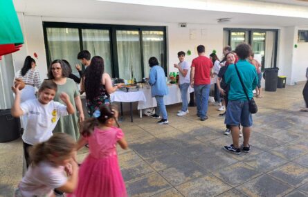 Várias pessoas – crianças, jovens e adultos – a dançar e conviver em espaço exterior. Outras pessoas, ao fundo da imagem, estão junto de mesas com bebidas e produtos alimentares.