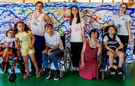 Grupo de oito pessoas, lado a lado, fotografadas em espaço interior. Ao fundo da imagem nota-se que alguém segura um cartaz gigante repleto de pinturas e traços coloridos.