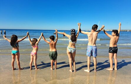 Seis crianças de costas, em pé, na praia (à beira-mar). Estão todas de mãos dadas e braços levantados. Ao fundo, no mar, mais algumas pessoas divertem-se.