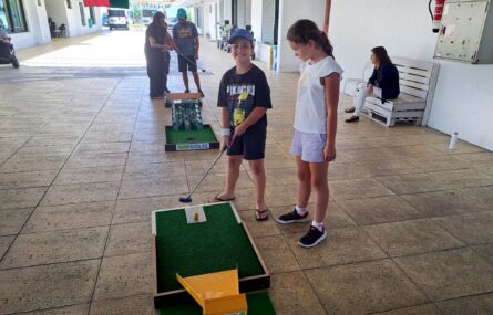 Duas crianças jogam mini-golfe. Mais atrás outros adultos e jovens desenvolvem a mesma atividade.