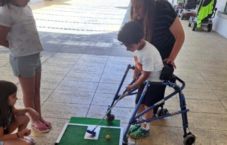 Uma criança joga mini-golfe. Ao lado um adulto acompanha a atividade e outras duas crianças assistem.