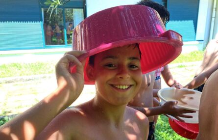 Uma sorridente criança coloca, na cabeça, um enorme recipiente de plástico. Atrás há mais crianças que também seguram recipientes similares.