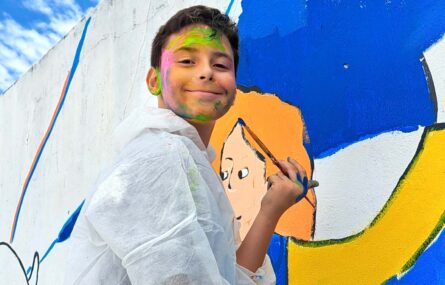 Uma criança segura na mão um pincel com o qual está a pintar um mural exterior. A criança tem o rosto pintado com traços de várias cores.