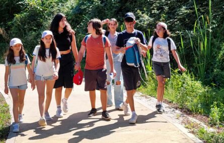 Sete crianças e jovens em percurso por caminho exterior rodeado de plantas e árvores.