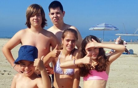 Fotografia realizada em areal, na praia. Em primeiro plano estão cinco crianças e jovens. Mais ao fundo o mar – onde outras pessoas tomam banho e se divertem.