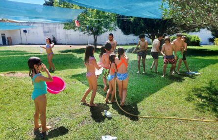 Mais de uma dezena de crianças a brincar em espaço relvado exterior. Estão a usar uma mangueira para lançar água enquanto outras seguram recipientes (também cheios de água).