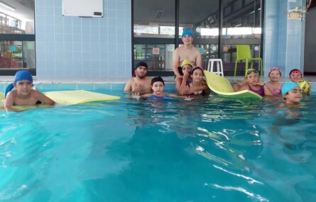 Fotografia realizada dentro de uma piscina interior na qual, dentro de água, estão 11 crianças e jovens.