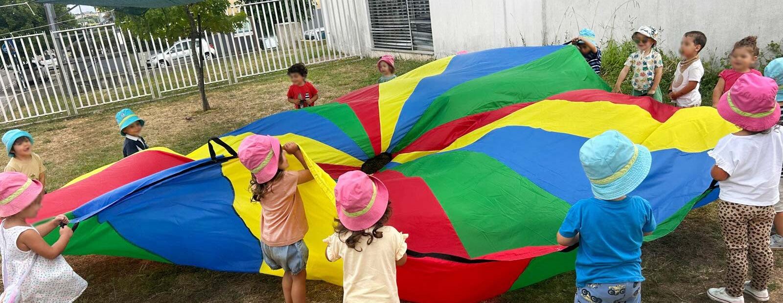 Grupo de mais de uma dezena de crianças em jardim exterior. Quase todas estão a segurar um enorme pano com várias cores. Os rostos de crianças visíveis e identificáveis foram digitalmente desfocados.