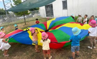Grupo de mais de uma dezena de crianças em jardim exterior. Quase todas estão a segurar um enorme pano com várias cores. Os rostos de crianças visíveis e identificáveis foram digitalmente desfocados.