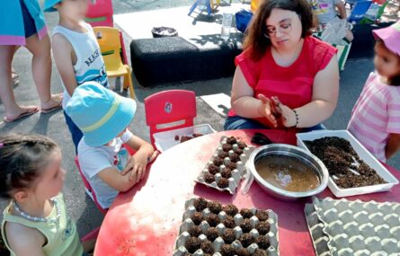 Várias crianças junto de uma mesa na qual um adulto, sentado, prepara o que aparentam ser brigadeiros de chocolate. Os rostos de crianças visíveis e identificáveis foram digitalmente desfocados.