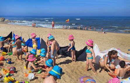 Perto de duas dezenas de crianças na praia. Mais ao fundo inúmeras pessoas a tomar banho no mar. Os rostos de crianças visíveis e identificáveis foram digitalmente desfocados.