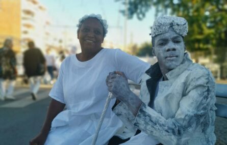 Duas pessoas, ambas vestidas de branco, sentadas num banco colocado numa rua.
