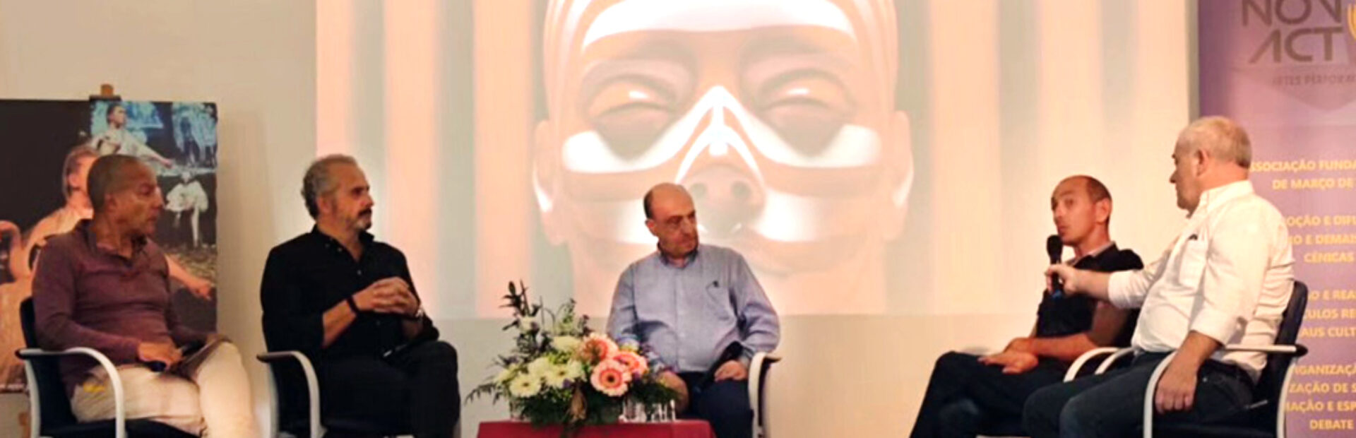 Cinco pessoas sentadas em palco. Uma das pessoas segura o microfone enquanto outra aparenta estar a falar. Ao centro uma pequena mesa com um arranjo floral. Em projeção multimédia, na parede ao fundo, imagem do cartaz do IV Fórum de Teatro Associativo e Artes Performativas.