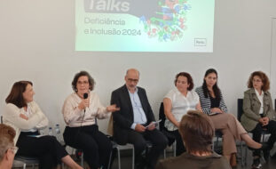 Seis pessoas sentadas junto de uma parede na qual está a ser projetada uma imagem multimédia. Uma das pessoas segura um microfone enquanto gesticula e aparenta estar a falar.