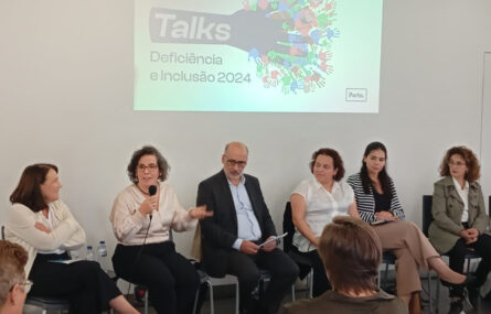 Seis pessoas sentadas junto de uma parede na qual está a ser projetada uma imagem multimédia. Uma das pessoas segura um microfone enquanto gesticula e aparenta estar a falar.
