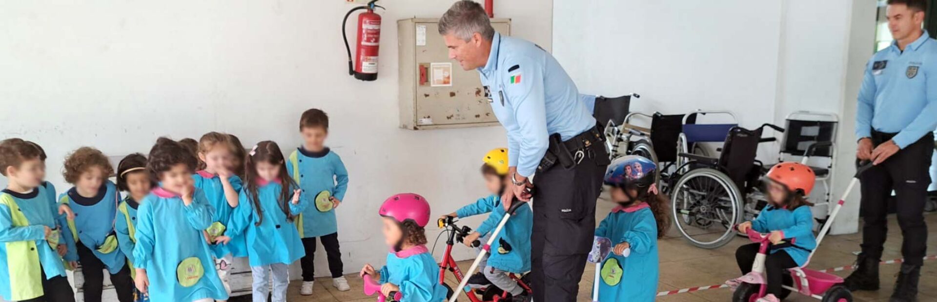 Grupo de perto de uma dezena de crianças. Junto das crianças estão dois adultos (com farda da PSP). As crianças experimentam com triciclos e trotinetas fazer percursos delimitados no chão. Os rostos visíveis das crianças foram digitalmente desfocados.