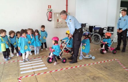 Grupo de perto de uma dezena de crianças. Junto das crianças estão dois adultos (com farda da PSP). As crianças experimentam com triciclos e trotinetas fazer percursos delimitados no chão. Os rostos visíveis das crianças foram digitalmente desfocados.