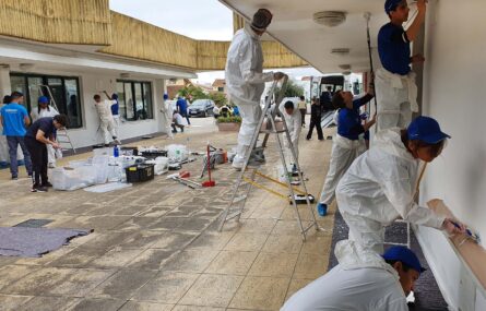 Em espaço exterior de unidade da APPC, inúmeras pessoas pintam e fazem pequenos trabalhos de recuperação de paredes. Imenso material de trabalho espalhado no chão.