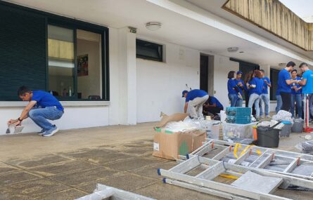 Material de pintura e trabalhos de recuperação espalhado no chão. Ao fundo, junto da parede do edifício, uma dezena de pessoas a conversar e a trabalhar.