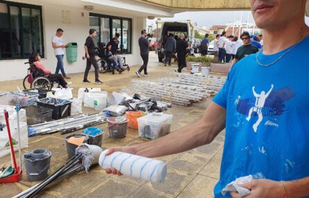 Várias pessoas transitam em espaço exterior repleto de material de pintura e de pequenas obras (espalhado no chão). Em primeiro plano uma pessoa com uma camisola azul com a inscrição “Just a change”.