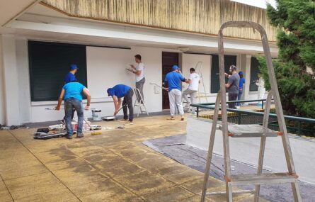 Nove pessoas em pintura de paredes de edifício. Uma das pessoas filma as restantes. Diverso material de pintura espalhado no chão.