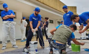 Cerca de uma dezena de pessoas com inúmeros materiais de pintura e construção. Enquanto uns preparam os materiais, outros estão a assistir.
