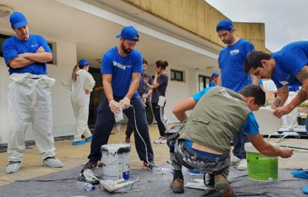 Cerca de uma dezena de pessoas com inúmeros materiais de pintura e construção. Enquanto uns preparam os materiais, outros estão a assistir.