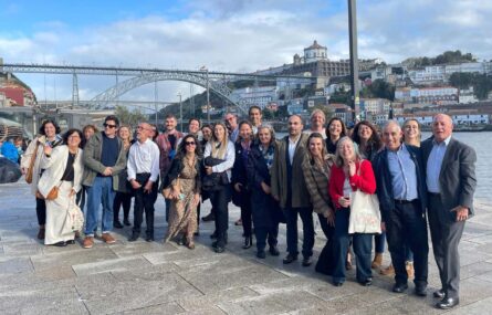 Perto de três dezenas de pessoas alinhadas para fotografia de grupo. Fotografia realizada em espaço exterior, na Ribeira do Porto, tendo como enquadramento uma ponte e vários edifícios em ambos os lados do rio (que se vislumbra atrás do grupo).