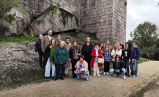Várias pessoas em passadiço de metal no que aparenta ser zona de muralhas de um castelo. Em primeiro plano, de costas, uma pessoa usa o telemóvel para fotografar as restantes.