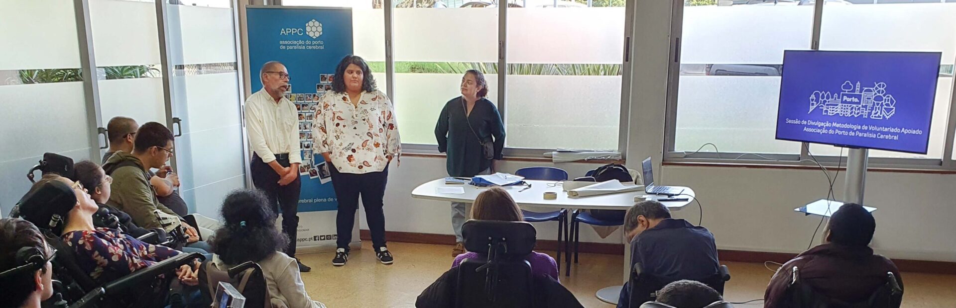 Grupo de pessoas, de costas, ouve o que três pessoas, em pé, estarão a dizer. Mesa com diverso material pousado. Cartaz da APPC do lado esquerdo e ecrã com projeção do lado direito.