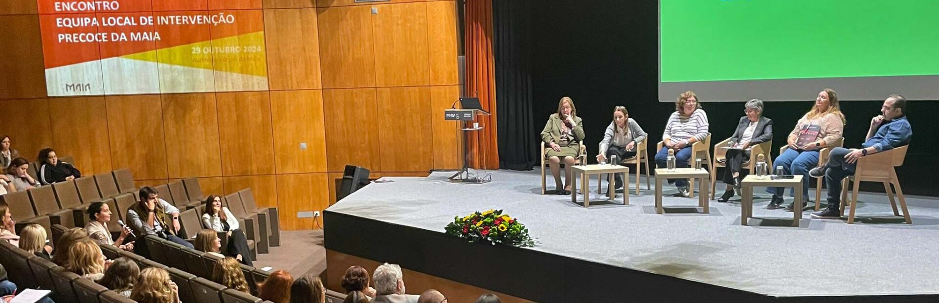 Fotografia de auditório amplo. Na zona do público várias dezenas de pessoas sentadas. No palco, igualmente sentadas (junto de três pequenas mesas) estão seis pessoas.
