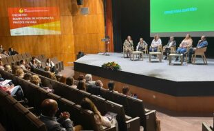 Fotografia de auditório amplo. Na zona do público várias dezenas de pessoas sentadas. No palco, igualmente sentadas (junto de três pequenas mesas) estão seis pessoas.